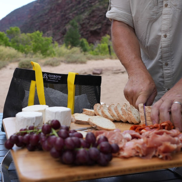 Person preparing a charcuterie board outdoors with a scenic background and Oxbow Supply Market Bag